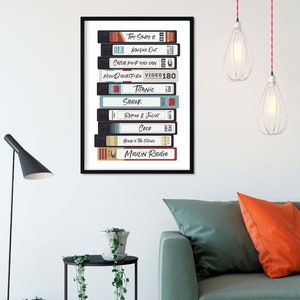 Może przedstawiać: Oprawiony plakat przedstawiający stos kaset VHS z tytułami film&oacute;w, takimi jak "Toy Story 2", "Knives Out" i "Titanic". Kasety są w r&oacute;żnych kolorach, z tytułami w białej i czerwonej czcionce. Plakat jest zawieszony na białej ścianie.