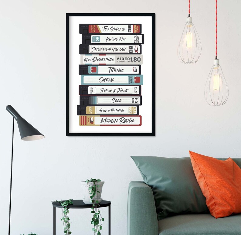 Może przedstawiać: Oprawiony plakat przedstawiający stos kaset VHS z tytułami film&oacute;w, takimi jak "Toy Story 2", "Knives Out" i "Titanic". Kasety są w r&oacute;żnych kolorach, z tytułami w białej i czerwonej czcionce. Plakat jest zawieszony na białej ścianie.