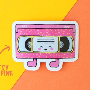 May include: A pink and yellow retro VHS cassette tape sticker with a smiling face and legs. The sticker has a 90s style pattern.