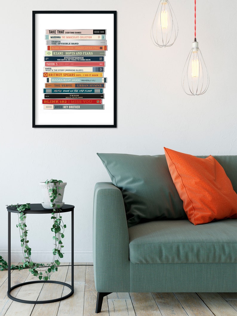May include: A framed print of a stack of album covers, featuring artists like Madonna, Take That, Maroon 5, and Britney Spears. The album covers are arranged in a vertical stack, with the titles and artist names visible. The print is framed in a black frame and is set against a white background.