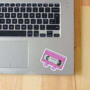 May include: A pink and yellow cassette tape with a smiling face and small legs. The cassette tape is on a silver laptop.