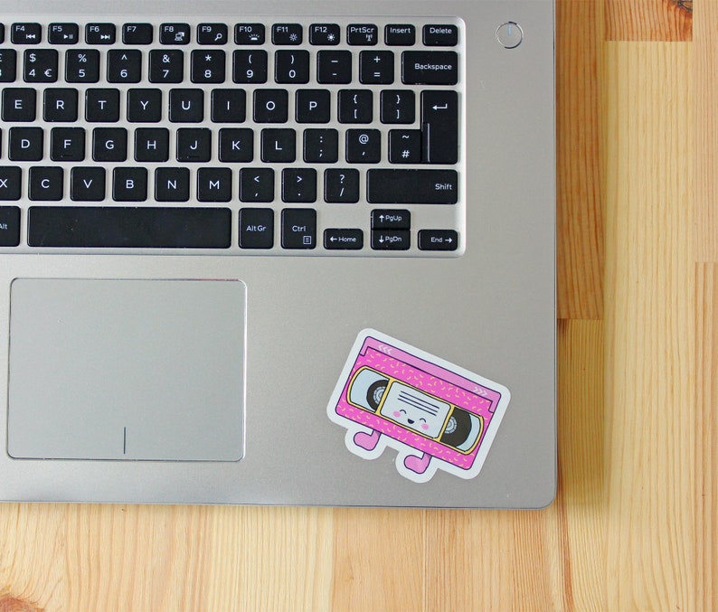 May include: A pink and yellow cassette tape with a smiling face and small legs. The cassette tape is on a silver laptop.