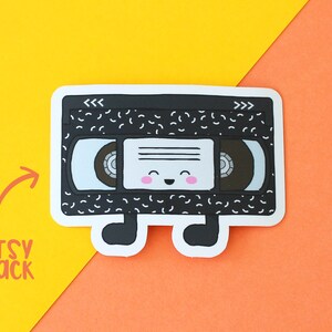 May include: A black and white vinyl cassette tape sticker with a smiling face and the words "<<<>>>" on the top. The sticker has a white background and is on a yellow and orange surface.