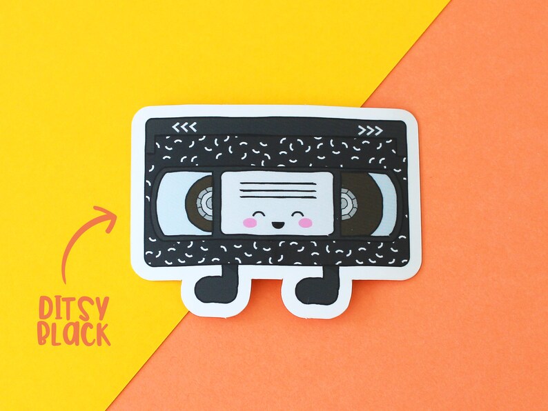 May include: A black and white vinyl cassette tape sticker with a smiling face and the words "<<<>>>" on the top. The sticker has a white background and is on a yellow and orange surface.