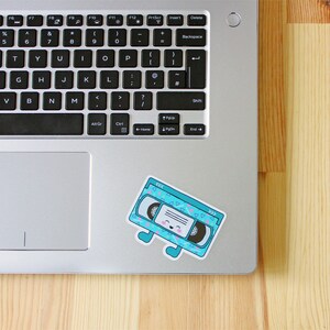 May include: A blue and white patterned VHS tape sticker with a smiling face and legs. The sticker is on a silver laptop.