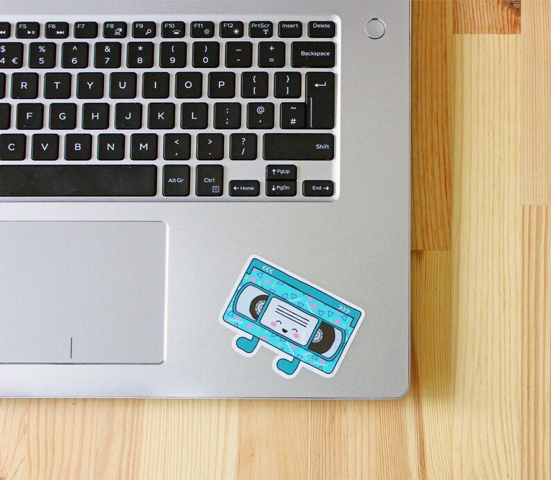 May include: A blue and white patterned VHS tape sticker with a smiling face and legs. The sticker is on a silver laptop.