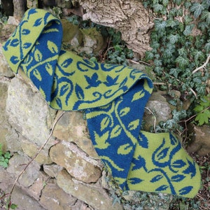 May include: A green and blue knitted scarf with a leafy pattern. The scarf is folded and draped over a stone wall.