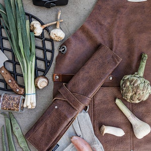 May include: A brown leather apron with the text "Brickwalls and Barricades" is displayed with various cooking ingredients. The ingredients include mushrooms, garlic, green onions, and an artichoke. A rolled leather knife case and a gray linen towel are also present.
