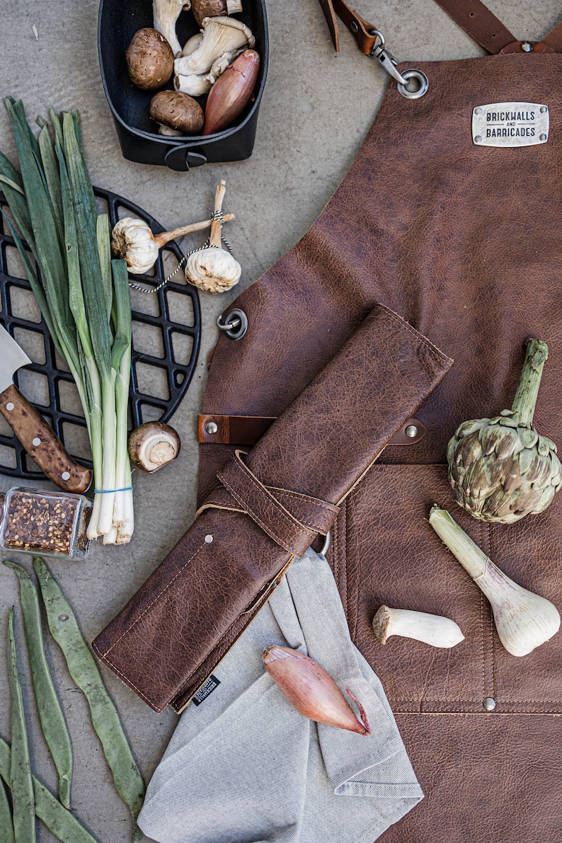 May include: A brown leather apron with the text "Brickwalls and Barricades" is displayed with various cooking ingredients. The ingredients include mushrooms, garlic, green onions, and an artichoke. A rolled leather knife case and a gray linen towel are also present.