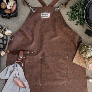 May include: A brown leather apron with the text "BRICKWALLS BARRICADES" on a metal tag. The apron is surrounded by cooking ingredients, including vegetables, a cast iron pot, and a cutting board with bread.