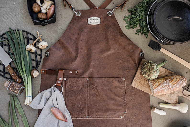 May include: A brown leather apron with the text "BRICKWALLS BARRICADES" on a metal tag. The apron is surrounded by cooking ingredients, including vegetables, a cast iron pot, and a cutting board with bread.