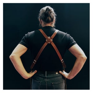 May include: Brown leather apron suspenders with a cross-back design. The suspenders feature adjustable straps and metal hardware. The leather is a warm, natural brown color, and the design is simple and functional. The suspenders are worn over a black t-shirt.