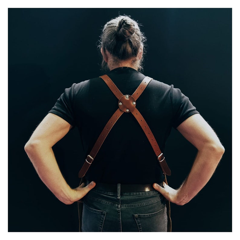 May include: Brown leather apron suspenders with a cross-back design. The suspenders feature adjustable straps and metal hardware. The leather is a warm, natural brown color, and the design is simple and functional. The suspenders are worn over a black t-shirt.