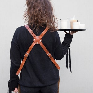 May include: A brown leather apron with a cross-back design, featuring adjustable straps and a central circular accent with rivets. The apron is worn over a black sweater. A black tray holds several cups of coffee.