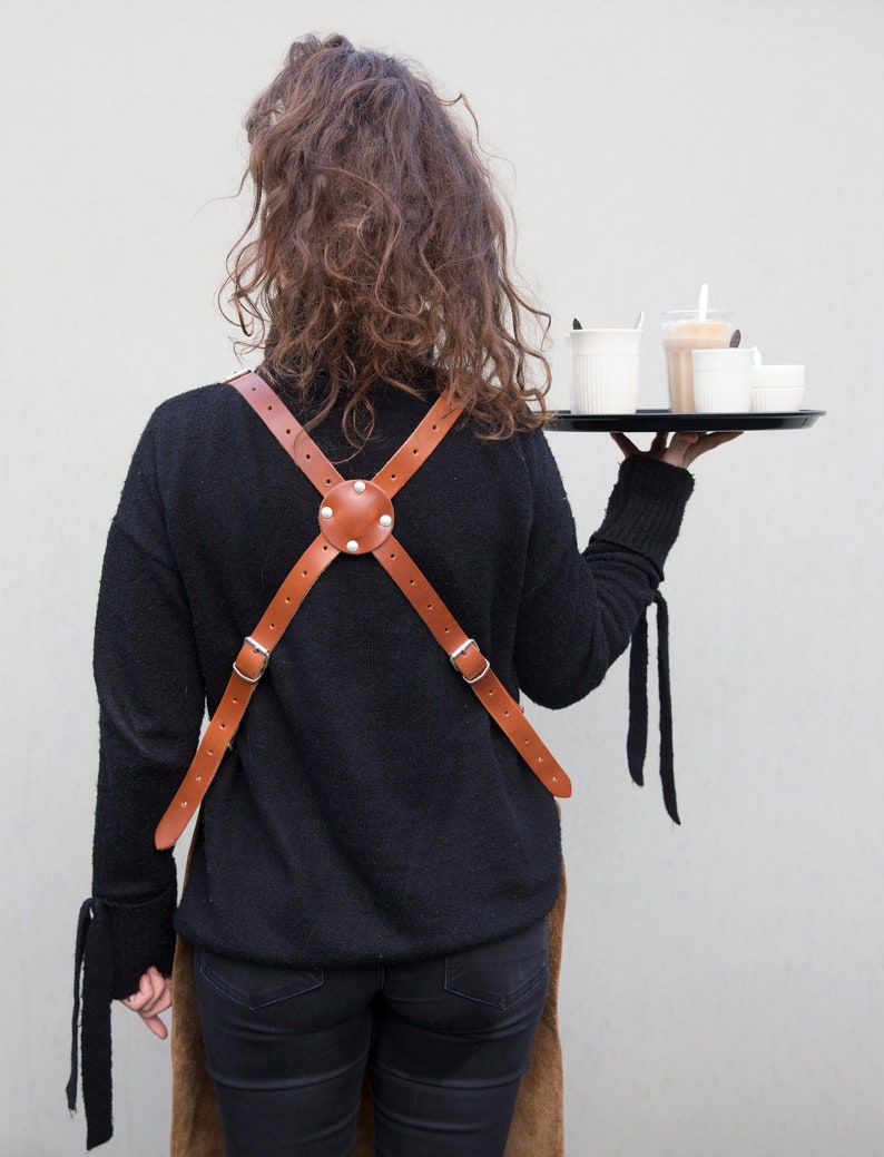 May include: A brown leather apron with a cross-back design, featuring adjustable straps and a central circular accent with rivets. The apron is worn over a black sweater. A black tray holds several cups of coffee.