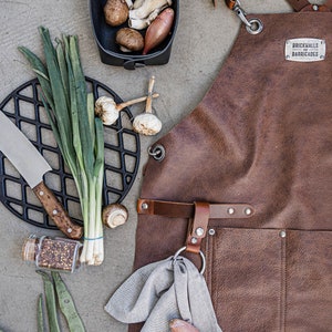 May include: A brown leather apron with the text "BRICKHALLS BARRICADES" is displayed with cooking ingredients. A black cast iron grill, a knife, mushrooms, garlic, and green onions are also visible.
