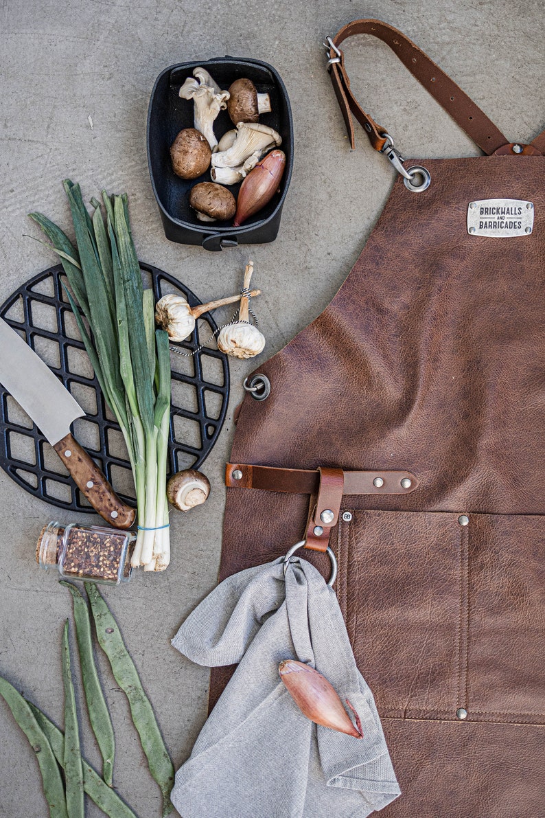 May include: A brown leather apron with the text "BRICKHALLS BARRICADES" is displayed with cooking ingredients. A black cast iron grill, a knife, mushrooms, garlic, and green onions are also visible.