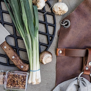 May include: Overhead shot of cooking and grilling essentials. Includes a brown leather apron, a black cast iron grill grate, fresh green onions, garlic bulbs, a knife, a mushroom, a jar of spices, and a gray cloth.