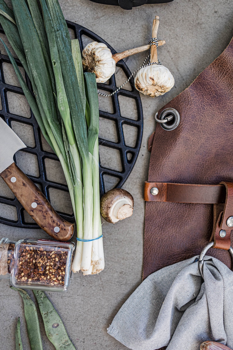 May include: Overhead shot of cooking and grilling essentials. Includes a brown leather apron, a black cast iron grill grate, fresh green onions, garlic bulbs, a knife, a mushroom, a jar of spices, and a gray cloth.