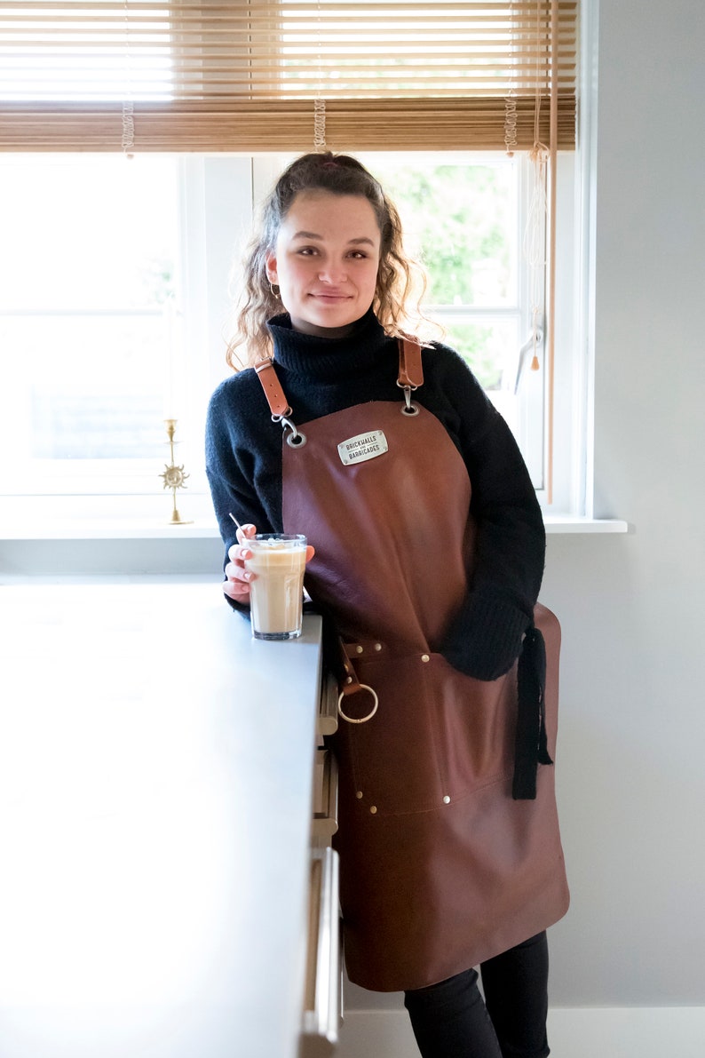 May include: A brown leather apron with adjustable straps and metal hardware. The apron has a pocket and a metal ring. The apron has the text "BRICKWALLS BARRICADES" on a small metal tag. The person is wearing a black turtleneck.