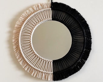 Puede incluir: Espejo redondo decorativo con diseño macramé. Dividido en dos mitades, beige natural y negro. Los flecos añaden textura. El espejo está colgado en una pared blanca. Diámetro aproximado de 40 cm.