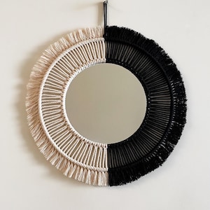 May include: A circular mirror with a macrame design, split into beige and black halves. The fringe detailing adds texture. The mirror is suspended on a white wall. The design is approximately 40 cm in diameter.
