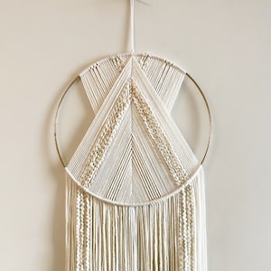 Puede incluir: Un tapiz de macramé blanco con un aro dorado y flecos largos. El macramé está tejido en un patrón geométrico.