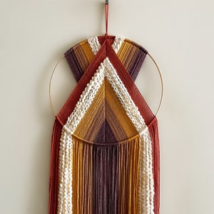 Pode incluir: Uma tapeçaria de macramé de parede com um aro circular dourado e um design geométrico em tons de vermelho, laranja, amarelo, marrom e branco.