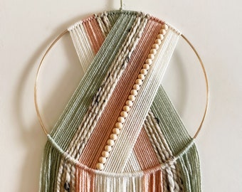 May include: A macrame wall hanging with a circular wooden frame and a fringe of yarn in shades of green, peach, and white. The macrame is decorated with wooden beads.