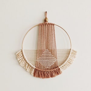 May include: A circular macrame wall hanging with a geometric design in shades of brown and cream. The hanging features a wooden bead at the top and fringe around the edges.