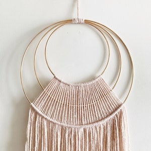 May include: A beige macrame wall hanging with three gold metal hoops. The hanging has a fringe of yarn at the bottom.