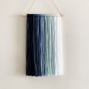 May include: A wall hanging made of yarn with a gradient of blue, green, and white colors. The yarn is attached to a wooden dowel rod and hangs from a string.