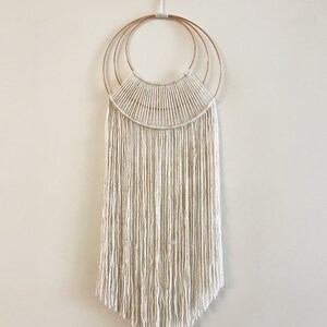 May include: A white macrame wall hanging with a copper ring design. The hanging has a fringe of white yarn.