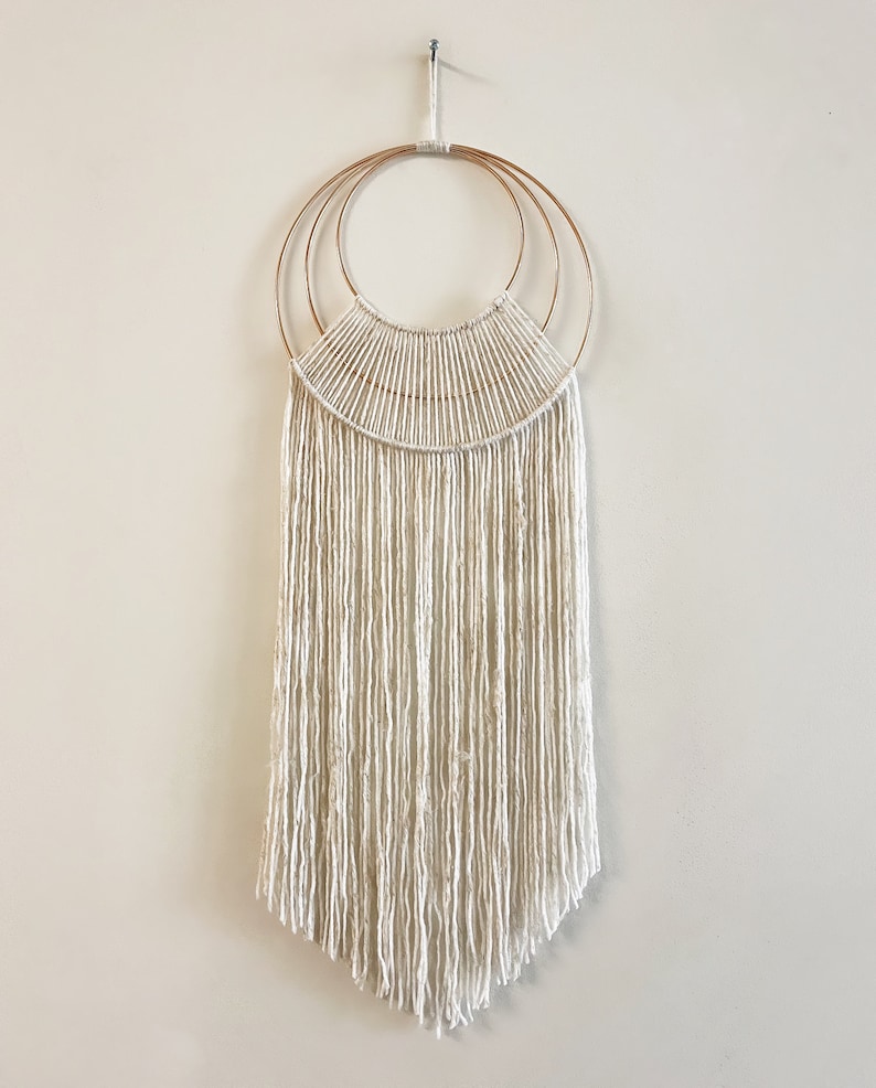 May include: A white macrame wall hanging with a copper ring design. The hanging has a fringe of white yarn.