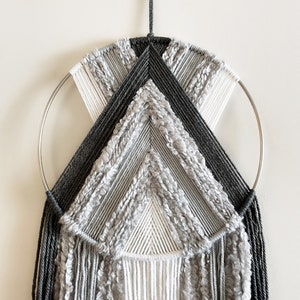 May include: A gray and white macrame wall hanging with a circular metal hoop. The hanging features a geometric design with fringe details.