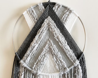 May include: A gray and white macrame wall hanging with a circular metal hoop. The hanging features a geometric design with fringe details.