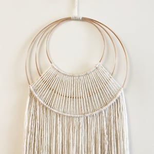 May include: A white macrame wall hanging with three concentric gold hoops. The macrame is made with thick white yarn and has a fringe at the bottom.