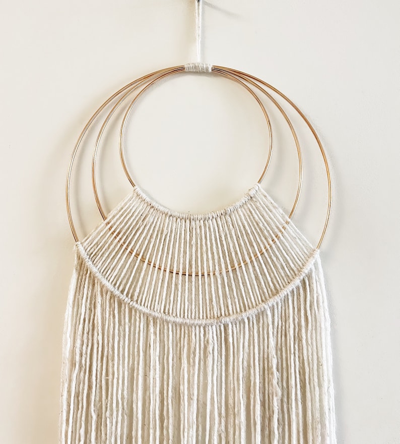 May include: A white macrame wall hanging with three concentric gold hoops. The macrame is made with thick white yarn and has a fringe at the bottom.