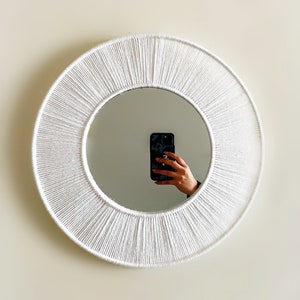 May include: A circular white mirror with a woven rope border. The mirror reflects a person holding a phone. The mirror is mounted on a light-coloured wall. Home decor.