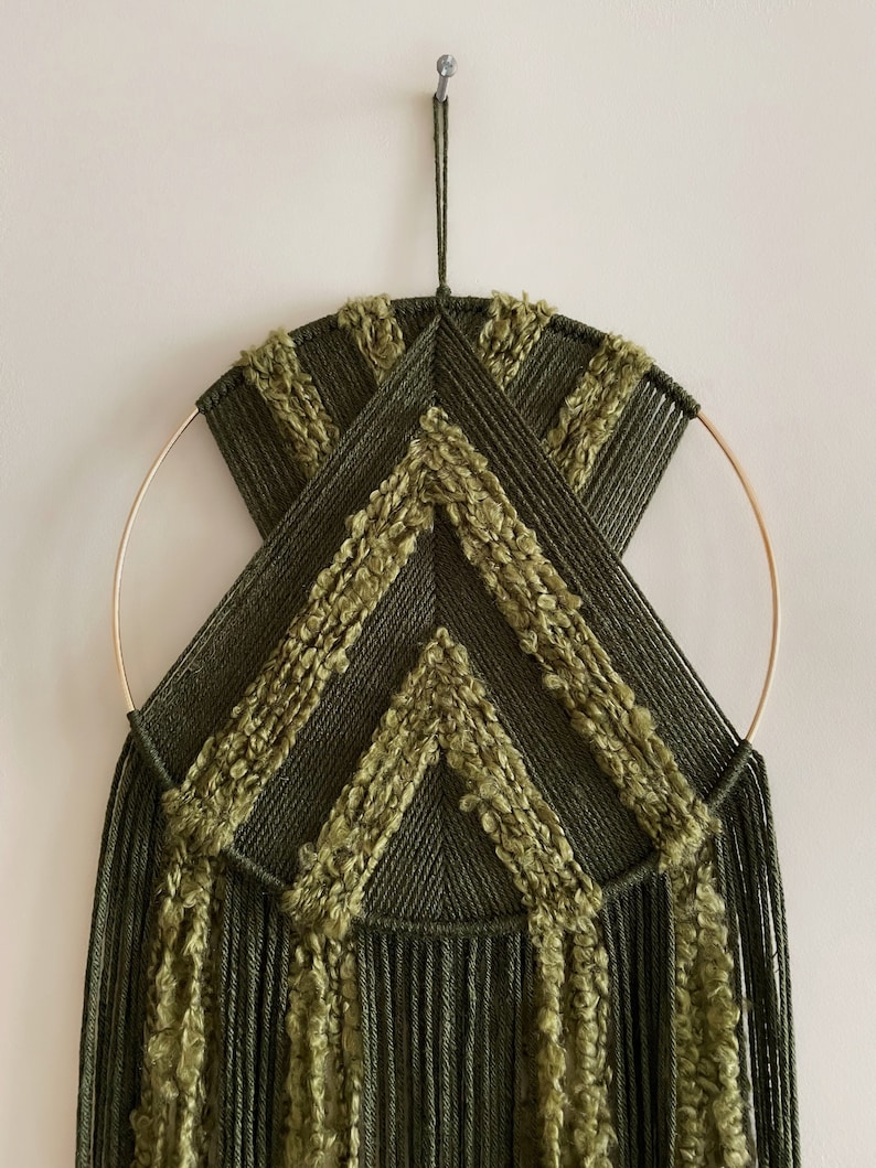 May include: A green and gold macrame wall hanging with a geometric design. The hanging features a circular hoop and long fringe.