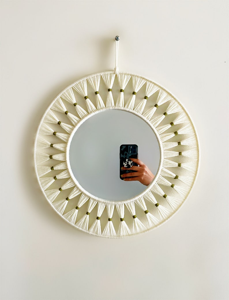 Op de afbeelding: Ronde witte spiegel met een macram&eacute;-frame met groene kwastjes. De spiegel hangt aan een witte muur.