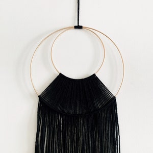 May include: Black macrame wall hanging with two gold hoops. The macrame is made with black yarn and has a fringe at the bottom.