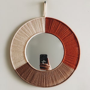 May include: A round mirror with a woven frame in shades of beige, brown, and orange. The frame is made of yarn and has a unique, textured design.