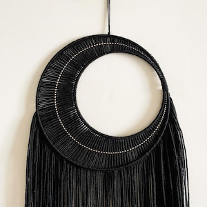 May include: A black macrame wall hanging in a crescent moon shape. The design features a woven, semi-circular top with a fringe of long, vertical strands. A thin, light-colored line of beads accents the top. The wall decor is suspended by a black cord.