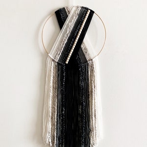May include: A black and white yarn wall hanging with wooden beads, woven into a circular design and hanging from a gold hoop.