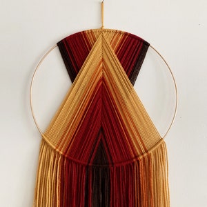 May include: A macrame wall hanging with a gold hoop and a geometric design in shades of brown, red, and yellow.