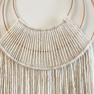 May include: A white macrame wall hanging with a gold metal hoop. The macrame is made with a thick white cord and has a fringe at the bottom.
