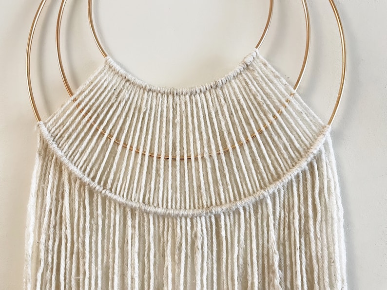 May include: A white macrame wall hanging with a gold metal hoop. The macrame is made with a thick white cord and has a fringe at the bottom.