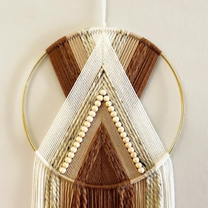 May include: A woven wall hanging with a gold metal ring, featuring a design in brown, cream, and white. The geometric pattern includes a triangle shape with wooden beads, creating a modern, bohemian aesthetic. The diameter is approximately 30 cm.