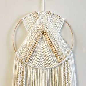 Puede incluir: Un tapiz de macramé blanco con un aro dorado y cuentas de madera. El tapiz tiene un diseño geométrico y flecos largos.